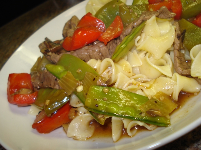 Beef with green and red peppers and snow peas on a bed of pasta on a white plate